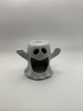 A small ghost-shaped ceramic holder or candle holder in light gray, featuring a hollow center for a cup or candle, with raised arms and a wide open mouth for a face.